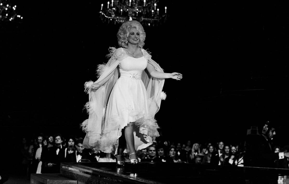 A black and white picture of Dolly Parton wearing a dress and walking across a stage.